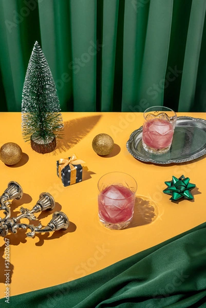 Fototapeta Vivid color retro futuristic Christmas dinner table setting with a tree, gift, yellow bow, red ice cold cocktails in crystal glass with emerald green curtain background. New Year's eve party aesthetic