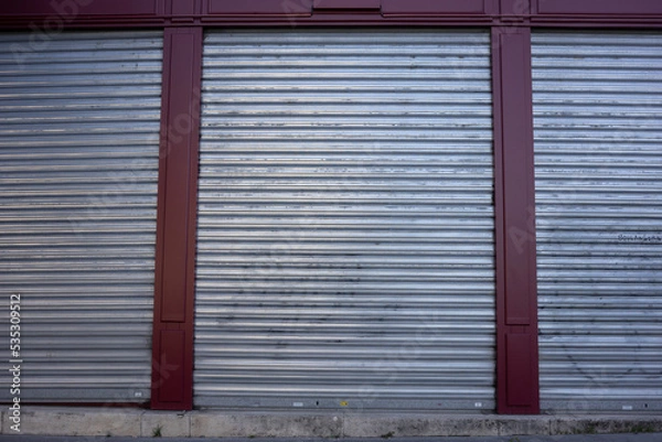 Fototapeta Three large silver metal gates are closed. Front view of a purple building with a sidewalk in the foreground. House facade in an industrial area