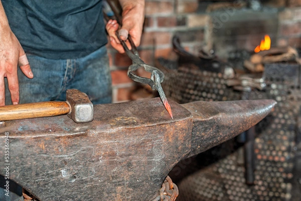Fototapeta A forger wearing blue jeans and a t-shirt holds fire thongs and a metal twisted bolt on a vintage anvil. There's rounding hammer tool with a wooden handle in the blacksmith foundry shop with a furnace