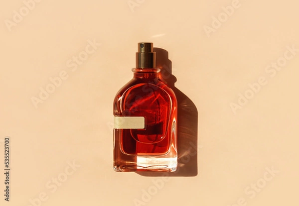 Fototapeta Transparent perfume bottle with red filling on a pastel beige background in the sunlight. Flat lay, top view. Luxury presentation mockup. Beautiful opened glass of aroma water