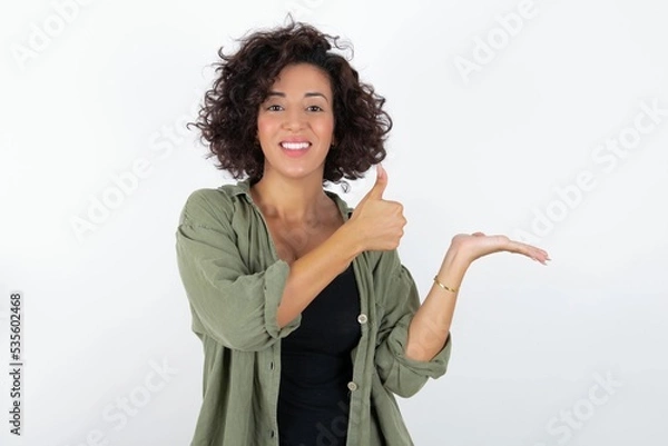 Fototapeta young beautiful woman with curly short hair wearing green overshirt over white wall Showing palm hand and doing ok gesture with thumbs up, smiling happy and cheerful.