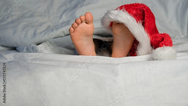 Fototapeta Children's heels lie on the bed under a white blanket, in a hat, Santa's cap. Concept of Christmas, happy holiday.