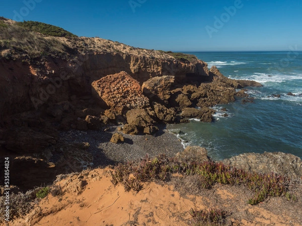 Obraz View of wild Rota Vicentina coast with ocean waves, sharp rock and green and red leaves of sour fig flower, Carpobrotus edulis near Vila Nova de Milfontes, Portugal. Sunny day, blue sky.