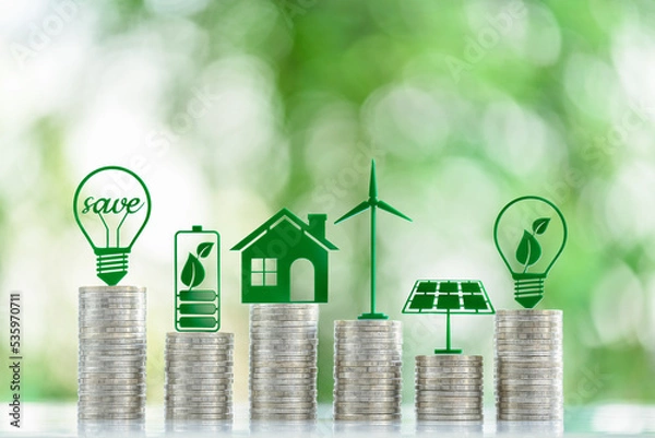 Fototapeta Renewable or clean energy generation prices and costs, financial concept : Green eco-friendly symbols atop coin stacks e.g. energy efficient light bulb, a battery, a solar cell panel, a wind turbine.