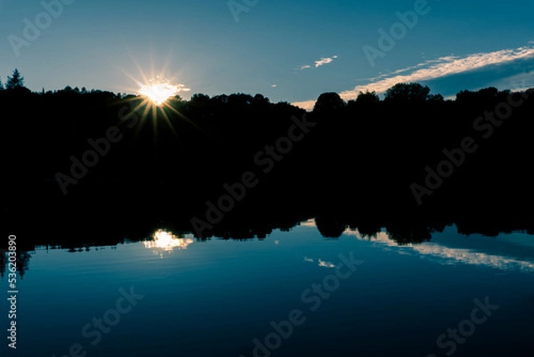 Fototapeta The sun sets in a glorious starburst behind silhouetted trees that line the edge of a lake.