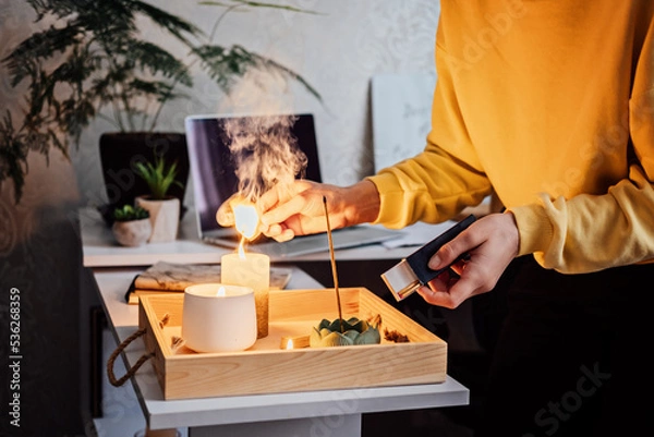 Fototapeta Meditation, relaxation after work. Woman getting ready meditating and lights up indian incense stick and candles. Woman hands with Smoke indian incense stick, burning candles on the table at home