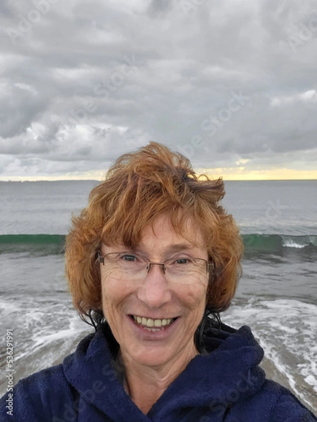 Obraz Mature woman taking a selfie after swimming in the sea. She is wearing a toweling robe to keep warm. Open or wild swimming in the sea is known to have benefits for physical and mental health. 