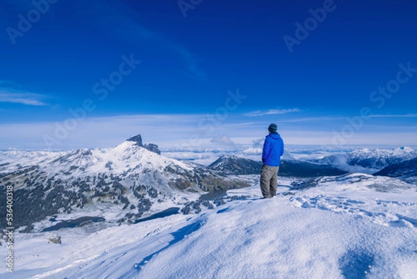 Fototapeta Adventurous athletic male hiker standing in snow at the summit of Panorama Ridge looking at The Black Tusk on a beautiful sunny day.
