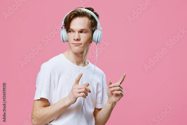 Fototapeta handsome, young man enjoying music in his headphones while standing on a pink background in a white t-shirt