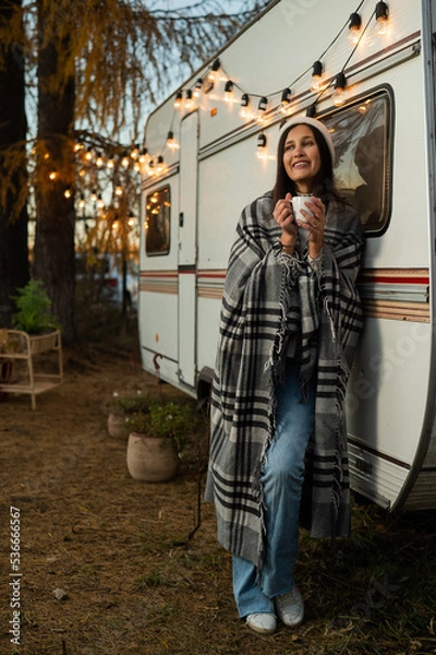 Fototapeta Caucasian woman in a knitted hat wrapped in a plaid and drinks a warming drink outdoors. Travel in a motor home in the fall.