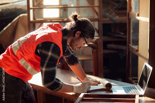 Obraz Young and handsome caucasian carpenter in the carpentry shop is working and checking customer's order in laptop to repair a furniture. small business and freelance craftsman.
