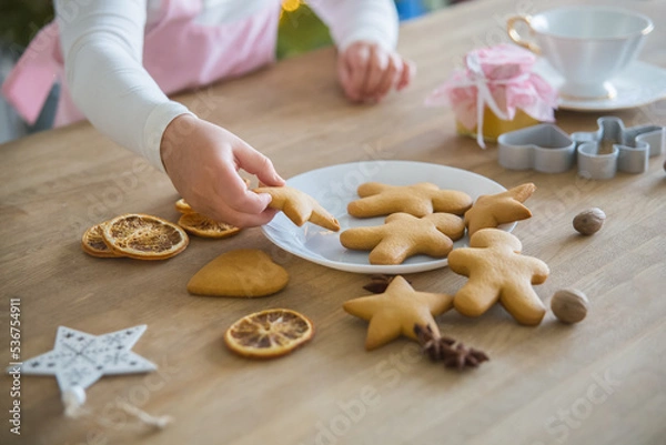 Fototapeta A child's hand takes from a white plate Christmas homemade cookies in the shape of a star, On a wooden table lie cookies gingerbread man, dried orange, anise, peenium molds and a white forfor cup