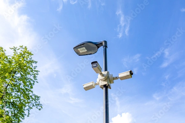 Fototapeta Modern lamppost and 3 surveillance cameras mounted in different directions in public park. Public space security systems