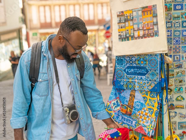 Fototapeta Young african american traveller man with cmera choosing the souvenirs at the typical souvenir shop. Travel in Portugal