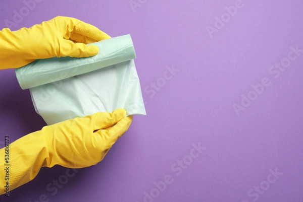 Obraz Janitor in rubber gloves holding roll of color garbage bags over violet background, top view. Space for text
