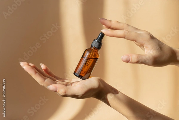 Obraz Bottle of serum in women's hands. Glass bottle with dropper cap in women's hands. Amber glass container with dropper lid for cosmetic products on brown background in sunlight