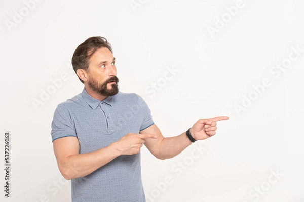 Fototapeta Bearded caucasian surprised man in blue t-shirt pointing fingers to the side at space for your presentation, stands on isolated white background.