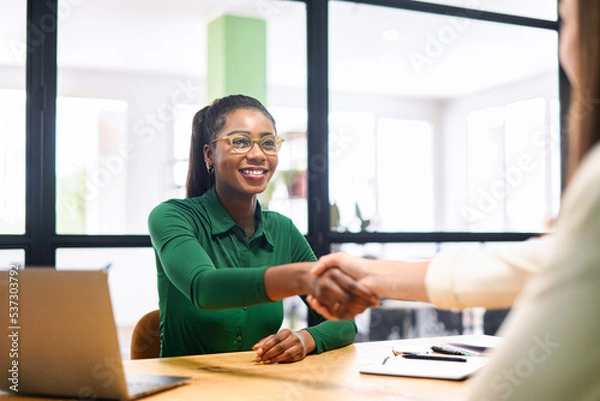 Obraz Happy female coworkers shaking hands in the office, african-american businesswoman smiling. Teamwork, partnerships and hiring concept