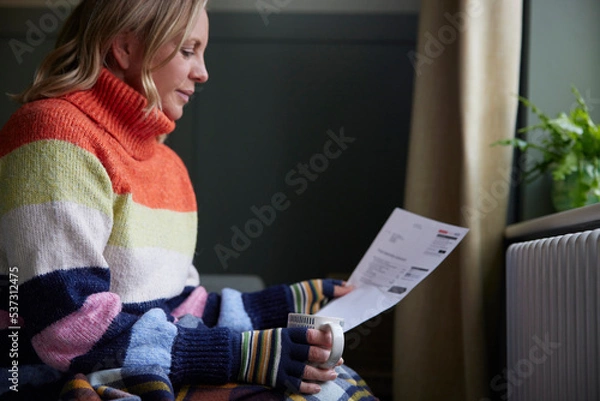 Obraz Woman In Gloves With Hot Drink And Bill Trying To Keep Warm By Radiator During Cost Of Living Energy Crisis
