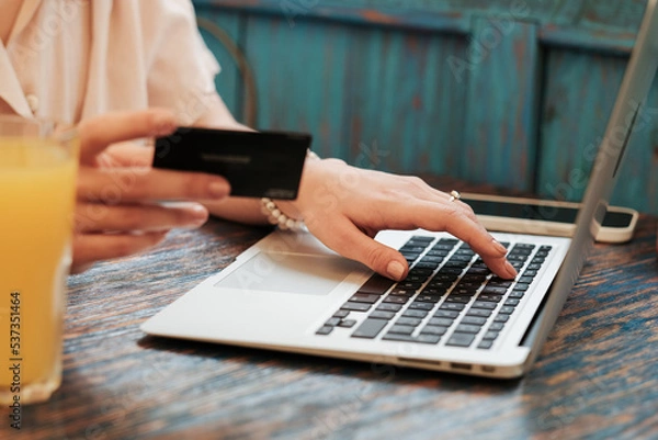 Fototapeta Close Up of Unrecognizable Woman Using Laptop for Online Shopping While Sitting in Restaurant, Female Customer Holding Credit Card and Making Payment Through the Internet.