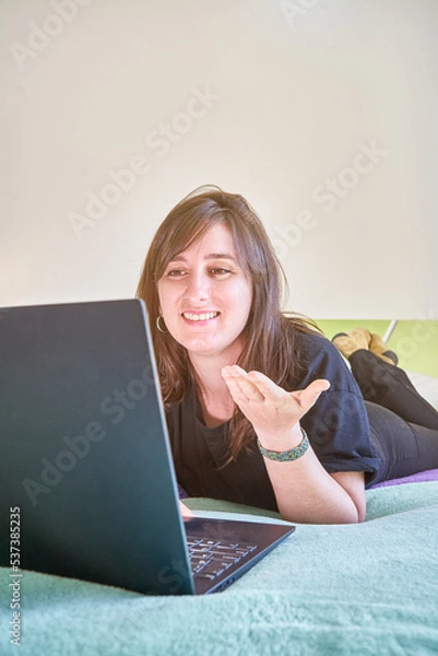 Fototapeta Vertical closeup of the woman with a laptop lying on the bed looking at the screen. Online chat.