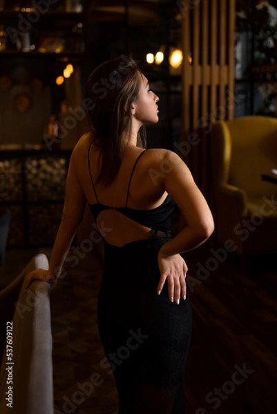 Obraz Young attractive woman in a black dress with her back open to the camera. Mysterious woman in thoughts in anticipation.
