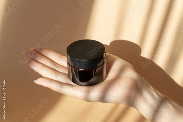 Obraz Amber jar with cream on woman's palm, on brown background in morning rays of sun. Concept of mockups, skin care