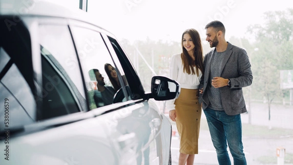 Fototapeta Attractive young couple confident bearded guy and his smiling pretty girlfriend are choosing new automobile together looking at luxury car in auto dealership and talking.