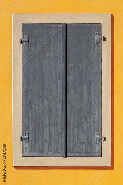 Fototapeta Rectangular window is covered with gray wooden shutters against a yellow wall. From the Window of the World series.