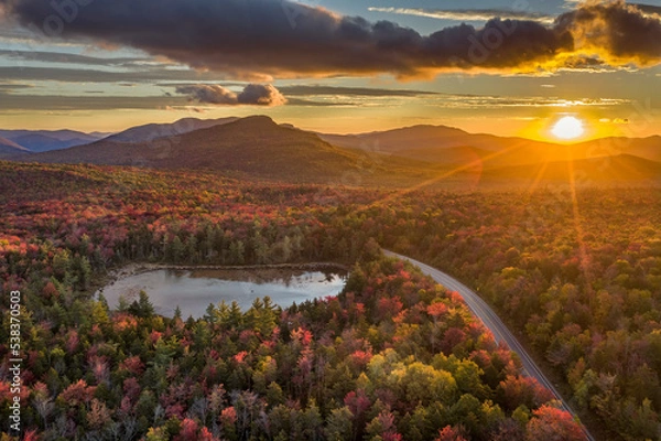 Fototapeta Sunrise over a scene of a road and lake through a forest with trees in fall colors of red, orange and yellow, Kancamagus Highway, White Mountains, New Hampshire