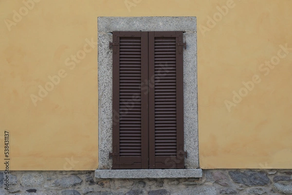 Fototapeta Window with brown wooden shutters on a yellow cement wall and grey stone brick wall, grey frame around the window with windowsill, 