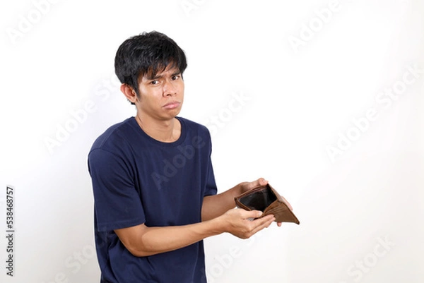 Fototapeta Disappointed young asian man standing while opening his empty wallet. Isolated on white background