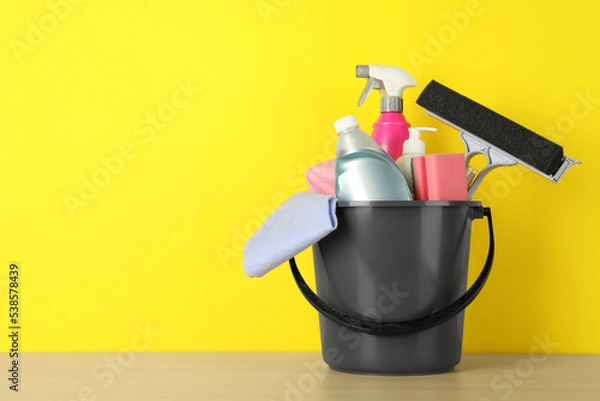 Fototapeta Bucket with different cleaning supplies on wooden floor near yellow wall. Space for text