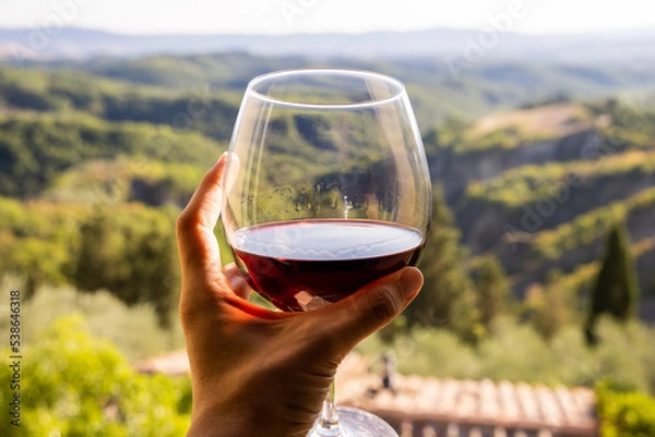 Fototapeta Woman holding a glass of red wine with beautiful landscape of Italy in a background on a sunny day. View from the window.