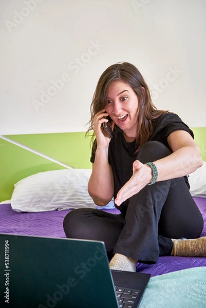 Fototapeta Vertical shot of a young woman sitting on a bed with a laptop and happily talking on a phone