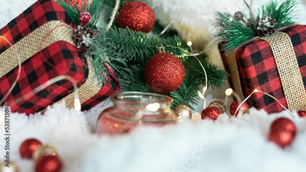 Obraz Decor from gifts in a checkered wrapper, branches of a Christmas tree, lights of a New Year's garland, a red candle, red and gold balls on a white fluffy background. Place for text. 