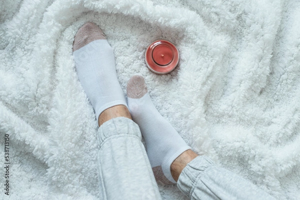 Obraz Women's legs in socks and warm gray pants on a white fluffy plaid, at home in a cozy autumn season. Cold, no heating.