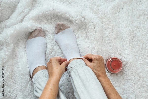 Obraz Women's hands put socks on legs sitting on a white fluffy plaid. Rest at home after a hard day's work. Varicose veins on legs. Swollen legs, problems with the bone in the legs.