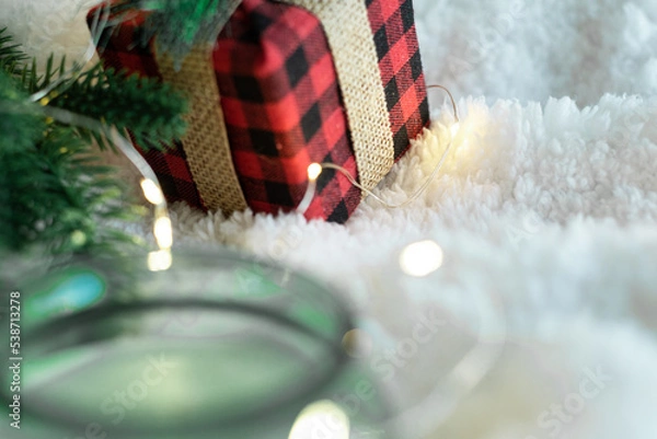 Obraz Focus on a gift in a plaid package decorated with a burlap ribbon, a warm-colored garland and a candle in a glass on a white soft background. Cozy holidays. New Year holidays 2023.