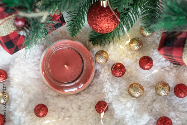 Obraz Red candle in glass and gifts in a checkered cloth wrapper, yellow lights on a garland, a sprig of a Christmas tree on a white soft background. Top view