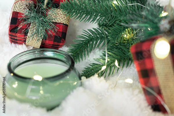 Obraz Green candle,gifts in a checkered cloth wrapper, yellow lights on a garland, a sprig of a Christmas tree on a white soft background. Focus on the gift. Postcard for the New Year and Christmas.