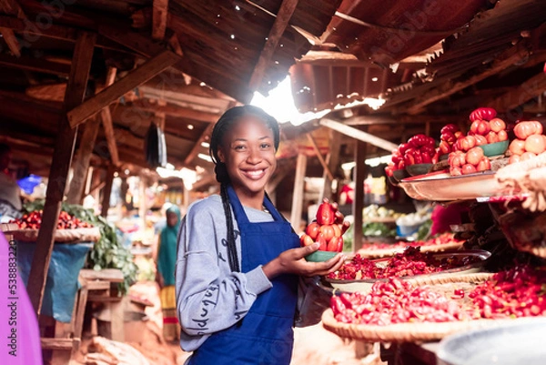 Obraz A african businesswoman wearing colourful Apron selling tomatoes and vegetables in a typical local african market