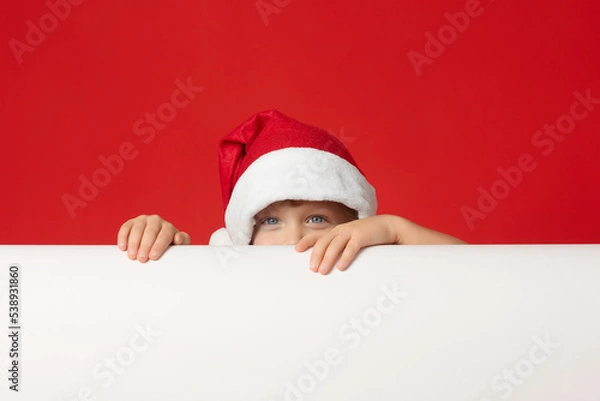 Fototapeta Sly look of little boy in red cap during the game, hiding behind white banner. Cute child in funny hat looks out from behind banner