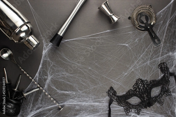 Fototapeta Various bar accessories such as: a shaker, muddler, strainer, as well as a bar spoon and a measuring stack lie against the backdrop of a spider web on a black matte table. Adult halloween celebration