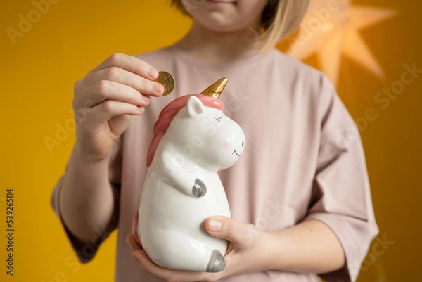 Obraz The girl holds a unicorn piggy bank in her hands and puts a coin in it.