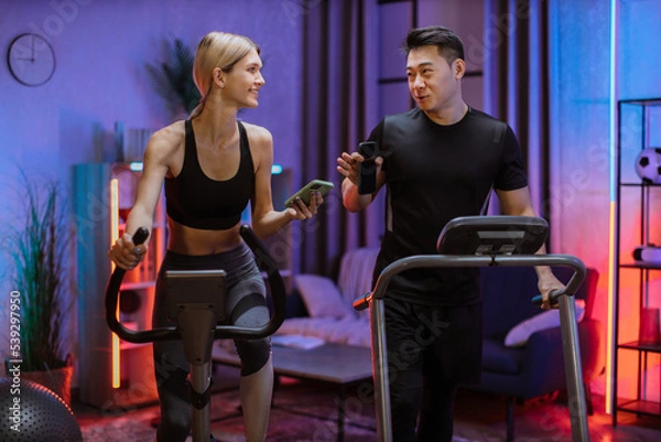 Fototapeta Blond caucasian sports women in sportswear cycling bike and fit asian man running on treadmill and showing each other photos from social networks.