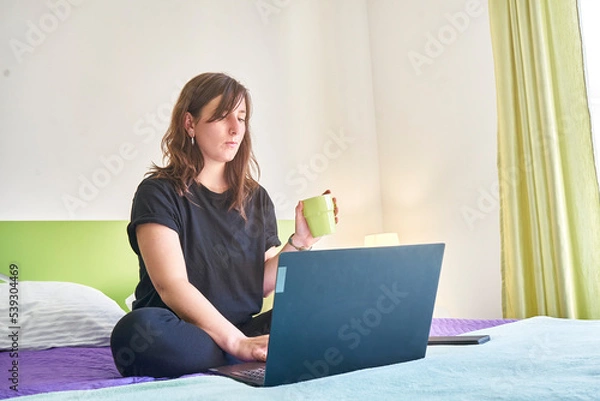Fototapeta Young Caucasian female sitting on the bed in front of a laptop with a cup in her hand
