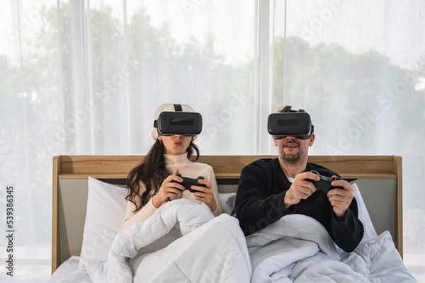 Fototapeta Caucasian couple wearing a VR headsets goggle device and holding game controller sitting on their bed playing game together.