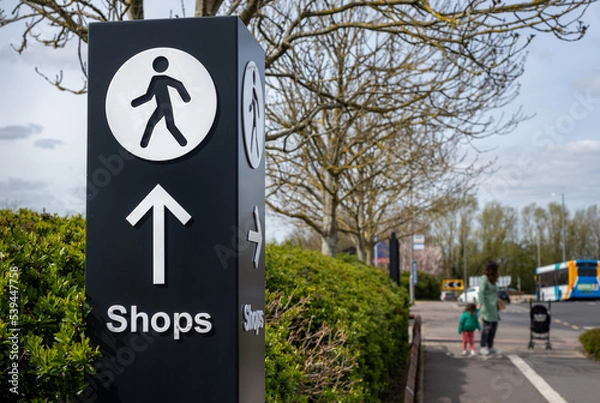 Fototapeta Large directional signage post with arrow and person walking icon pointing along path towards shopping retail area.