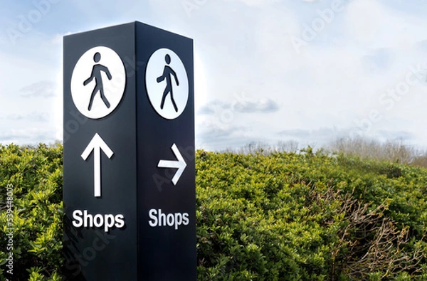 Fototapeta Black and white freestanding directional sign post with arrows and person symbol icon pointing towards shops. 